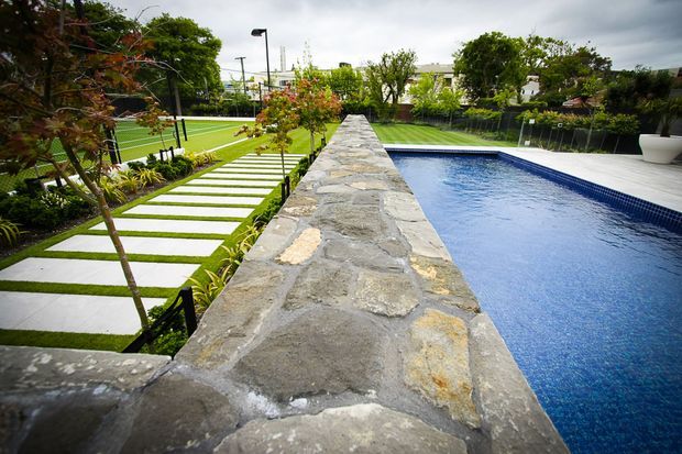 Landscaped outdoor recreation area with a long rectangular swimming pool beside a rough natural-stone masonry wall. A manicured lawn, large stepping-stone path, planted borders, fencing and modern outdoor lighting create a structured contemporary garden setting.
