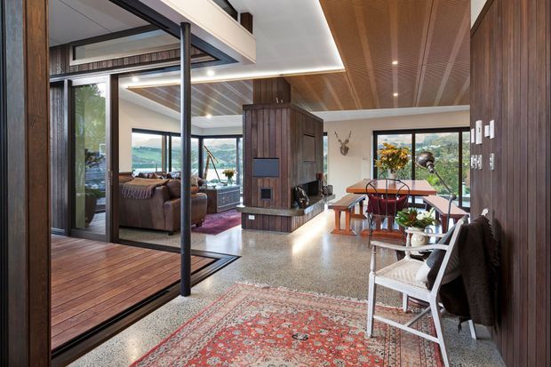 Open-plan mid-century-inspired interior with a dining area in the foreground, a living room beyond glazed sliding doors, and a kitchen or service zone behind a central timber-clad volume. Rich timber wall and ceiling lining, polished terrazzo flooring, extensive glazing, and a red patterned rug create a warm, refined connection to the surrounding landscape.