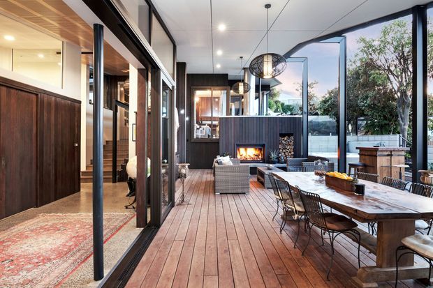 A sophisticated covered outdoor entertaining area with a timber deck, large dining table, lounge seating, and an outdoor fireplace set into a dark feature wall. Full-height glazed doors connect the deck to the interior, while a compact outdoor kitchen or bar area and surrounding greenery are visible at the far end. The space combines warm timber finishes with black metal framing and contemporary lighting.