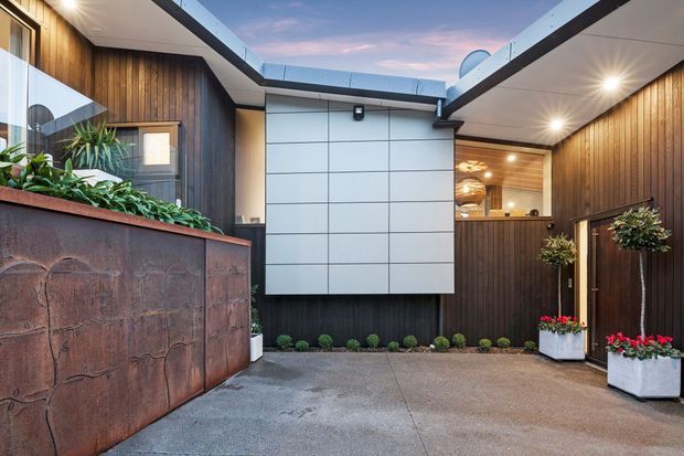 A contemporary courtyard entry with dark vertical timber cladding, a large light-panelled feature wall, recessed ceiling lighting, glazed roof openings, and a concrete-paved floor. A sculptural rust-toned planter wall and planted containers add texture and greenery, while a partially visible warm-lit interior lies beyond the entry.