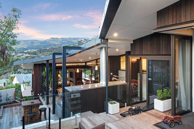 Elevated exterior view of a contemporary multi-level home on a sloping site, featuring dark vertical timber cladding, extensive glazing, slim metal framing, broad roof overhangs, and layered timber decks with glass balustrades. The open living area and kitchen are visible through the glazing, with a mountainous suburban landscape beyond.