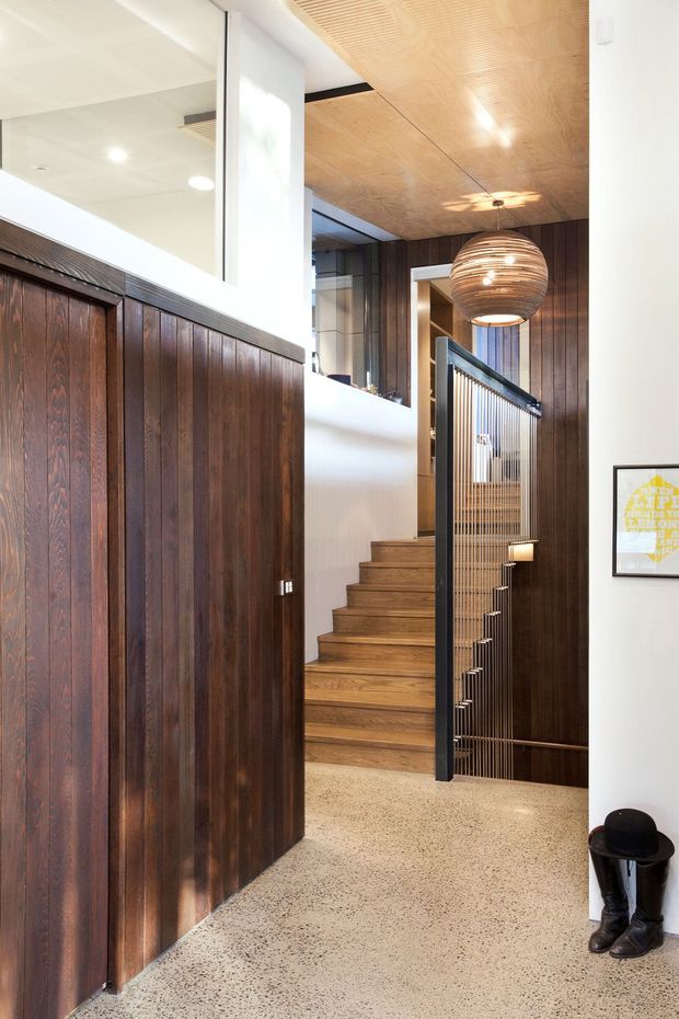 A refined residential interior hallway and stairway featuring dark vertical timber wall panelling, warm timber stair treads, a perforated timber ceiling, polished speckled concrete or terrazzo flooring, and a black metal stair balustrade with slender vertical rods. A large woven pendant light hangs above the stairwell, with white plaster walls, glazing, and framed artwork adding contrast.
