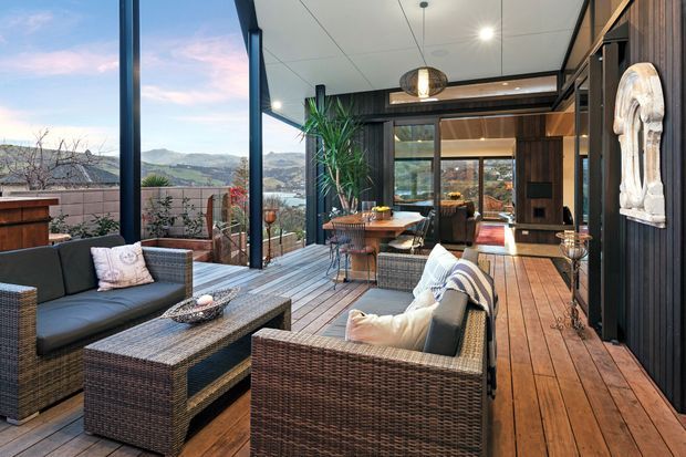A stylish enclosed outdoor living area with large glazed openings, a timber deck, woven outdoor sofas and coffee table, and a dining area. Dark timber wall cladding and structural framing contrast with the warm timber flooring, while a scenic mountainous landscape is visible through the glazing. The space feels like a sheltered outdoor lounge or sunroom.