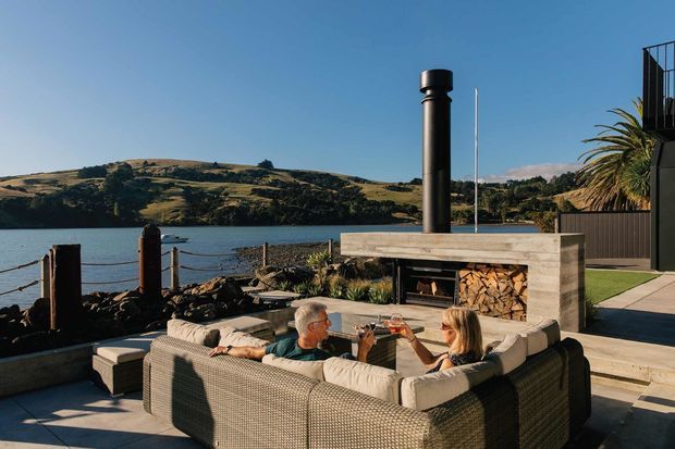 A waterfront outdoor living area with a large built-in fire pit and chimney, surrounded by a sectional sofa and low tables. The setting overlooks calm water and a hilly shoreline, with timber decking, stone edging, and a modern dark-clad building visible at right.