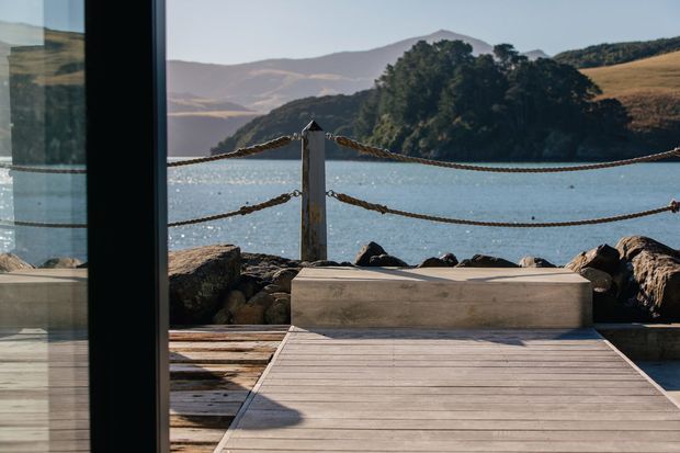 A timber deck or terrace viewed through full-height glazing, with a built-in concrete or stone bench, rope-and-post balustrade, rocky shoreline, and calm water backed by wooded hills and mountains. The scene has a refined coastal, outdoor-living character with warm natural materials and strong connection to the landscape.