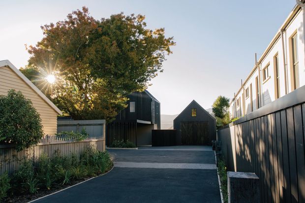 A suburban residential lane with a newly paved dark driveway leading between contemporary homes and a detached dark-clad gabled garage or studio. A light weatherboard house and planted garden border the left side, while a tall dark privacy fence and modern multi-level residence line the right. Mature trees and low late-afternoon sunlight create a warm, quiet streetscape.