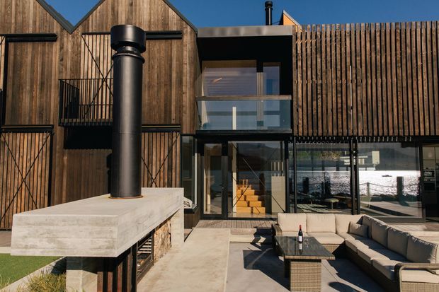 A contemporary multi-level residential exterior with a bold combination of vertical timber cladding, black metal framing, and extensive glazing. The courtyard terrace includes a large concrete outdoor fireplace, outdoor kitchen or bench structure, woven seating, and a low lounge arrangement. Gabled timber volumes contrast with a glazed central connection and upper balcony, creating a modern indoor-outdoor living area.