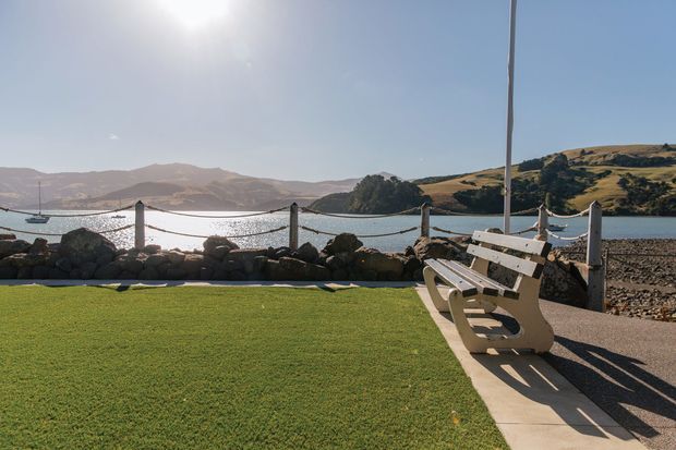 A sunny waterfront outdoor area with a broad lawn, a white timber bench, and a low stone-and-rope boundary overlooking a calm bay. Rolling hills and a small boat are visible across the water, creating an open coastal setting.
