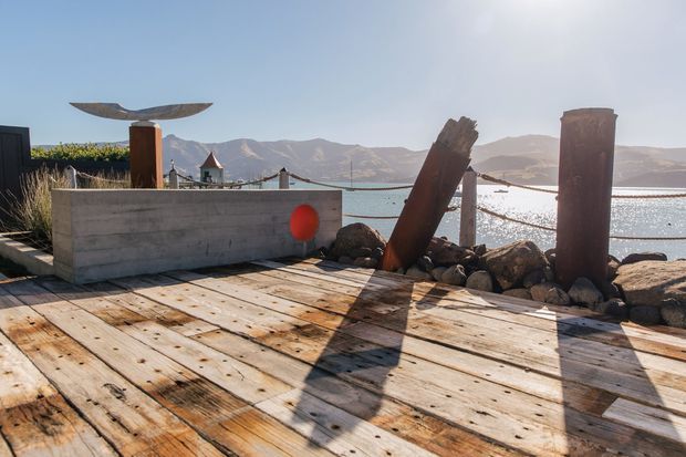 A sunlit timber deck or waterfront terrace overlooking a calm body of water and rugged mountain ranges. The deck features weathered wooden planks, a low concrete planter or wall, simple cable railing, large rocks along the shoreline, and sculptural rusted metal elements.