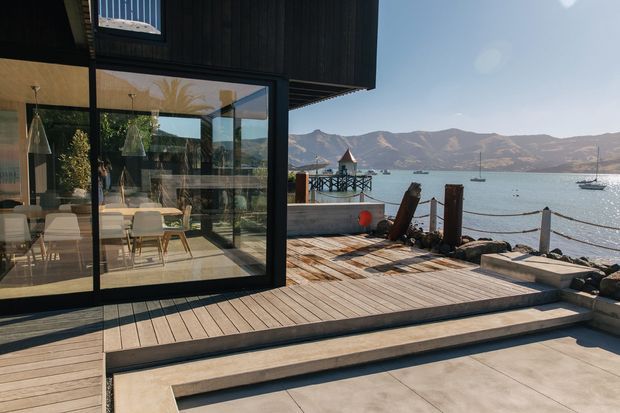 Modern waterfront home with a dark-clad glazed dining area opening onto a broad timber deck and pale paved terrace. The outdoor space includes steps, mooring posts and a small dock overlooking a calm bay with mountainous terrain beyond. The interior dining area is visible through extensive glazing.