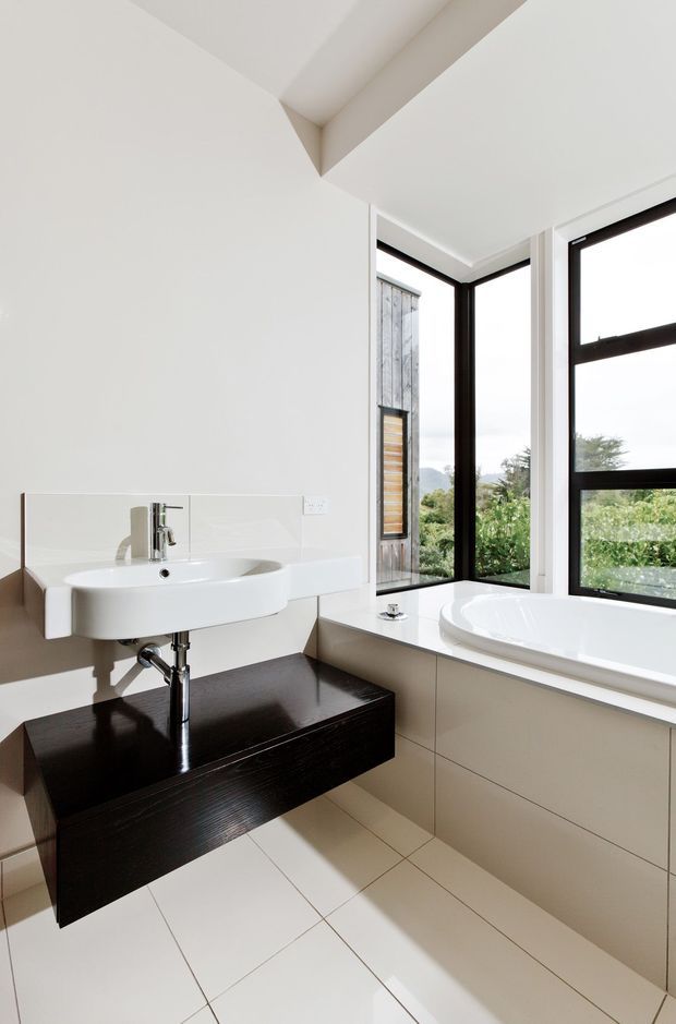 A bright, contemporary bathroom with a wall-mounted white basin and chrome tap above a dark timber vanity, a built-in white bathtub, pale tiled floor, light neutral cabinetry, and large black-framed corner windows overlooking greenery. Clean white walls and strong natural light create an airy, minimalist feel.