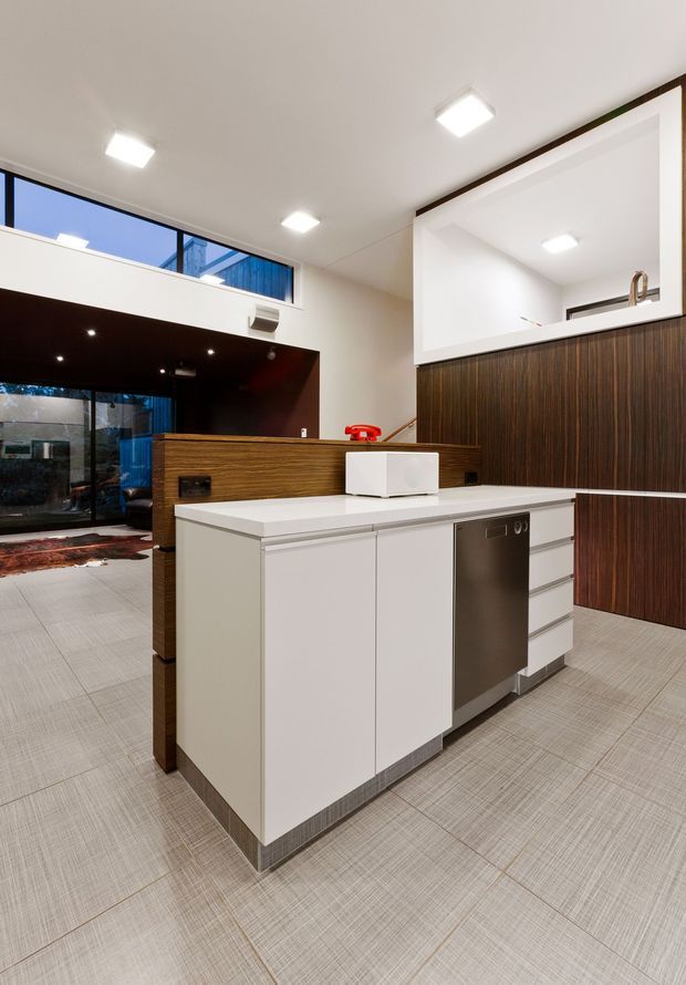 A contemporary open-plan kitchen with a compact white island, integrated dishwasher, pale tiled flooring, and dark wood-grain cabinetry. A raised timber breakfast bar separates the kitchen from a lower living area, with large glazing and a high-level window bringing in natural light. Recessed square ceiling lights and clean-lined finishes create a modern, functional interior.