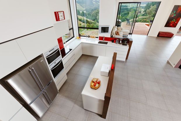 Bright contemporary open-plan kitchen viewed from above, with white handleless cabinetry, stainless-steel refrigerator and integrated ovens, a white island with timber seating, and large glazing opening toward a green landscape and outdoor deck. The kitchen flows into a spacious tiled living area.