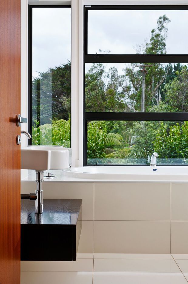 A bright, contemporary bathroom featuring a wall-mounted white basin on a dark timber vanity, a large white bathtub beneath broad black-framed windows, pale built-in cabinetry, and light tiled flooring. Lush greenery is visible outside through the glazing.