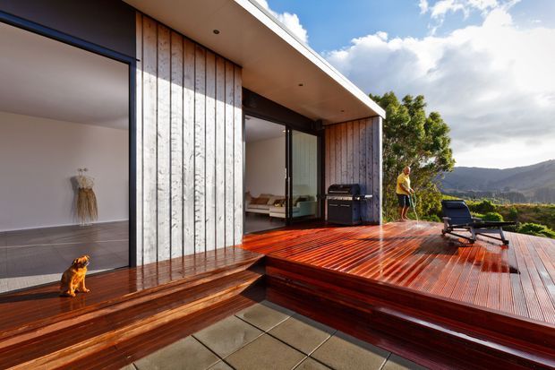Contemporary single-level home opening onto a raised timber deck, with vertical weathered timber cladding, expansive dark-framed glazing, integrated timber seating, and an outdoor entertaining area with a barbecue and loungers. A mountain landscape is visible beyond the deck.