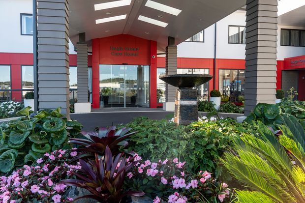 Exterior view of a contemporary commercial or institutional building entrance, with a broad covered canopy supported by layered rectangular columns. The entrance has vivid red wall panels, extensive glazing, clerestory-style windows, and a central tiered stone fountain. Lush landscaped planting with flowering plants and palms fills the foreground.