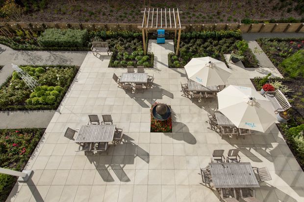 Aerial view of a landscaped outdoor courtyard or terrace paved with large light-coloured square slabs. Several weathered timber dining tables and chairs are arranged beneath large white umbrellas, with a central circular planter or feature and extensive raised garden beds around the perimeter. A timber pergola-like entry structure and planted retaining wall appear at the far edge.