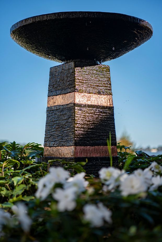 Close-up outdoor photograph of a dark, textured stone or concrete water feature resembling a broad mushroom-shaped fountain, mounted on a rectangular pedestal with horizontal copper-toned bands. It is surrounded by leafy planting and white flowers, with a blue sky and blurred landscape in the background.