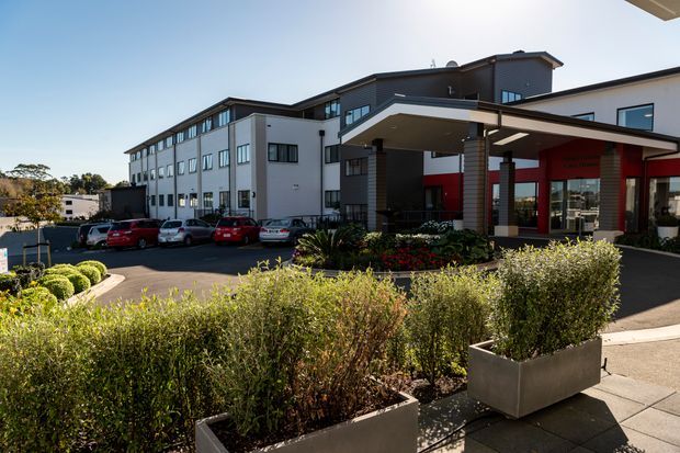 Exterior view of a contemporary multi-storey commercial or institutional building with a prominent covered entrance canopy, dark and light facade sections, extensive glazing, landscaped planter boxes, paved forecourt, and parked cars. The image is photographed from outside in bright daylight.