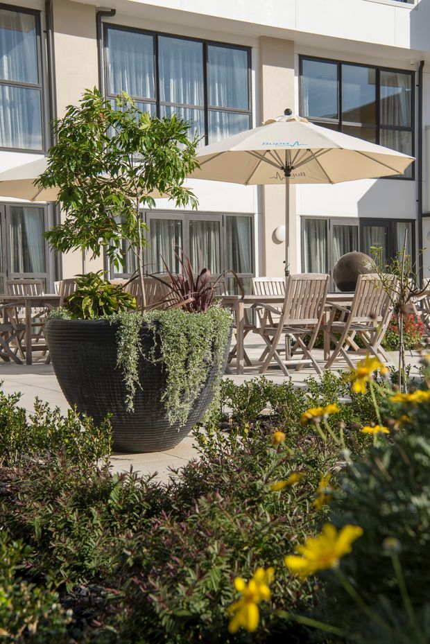 A sunny landscaped courtyard or terrace with abundant planting, large black textured planters, outdoor timber dining furniture, and cream patio umbrellas. The space fronts a contemporary multi-storey building with white rendered walls, dark-framed floor-to-ceiling windows, and glazed doors. The setting feels like a refined communal outdoor hospitality or residential courtyard.