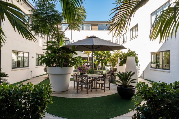A bright landscaped courtyard enclosed by white contemporary buildings, with tropical planting, a large central umbrella, outdoor dining furniture, potted plants, and artificial turf over a paved surface. The courtyard has an intimate resort-like atmosphere and visible surrounding windows and balconies.