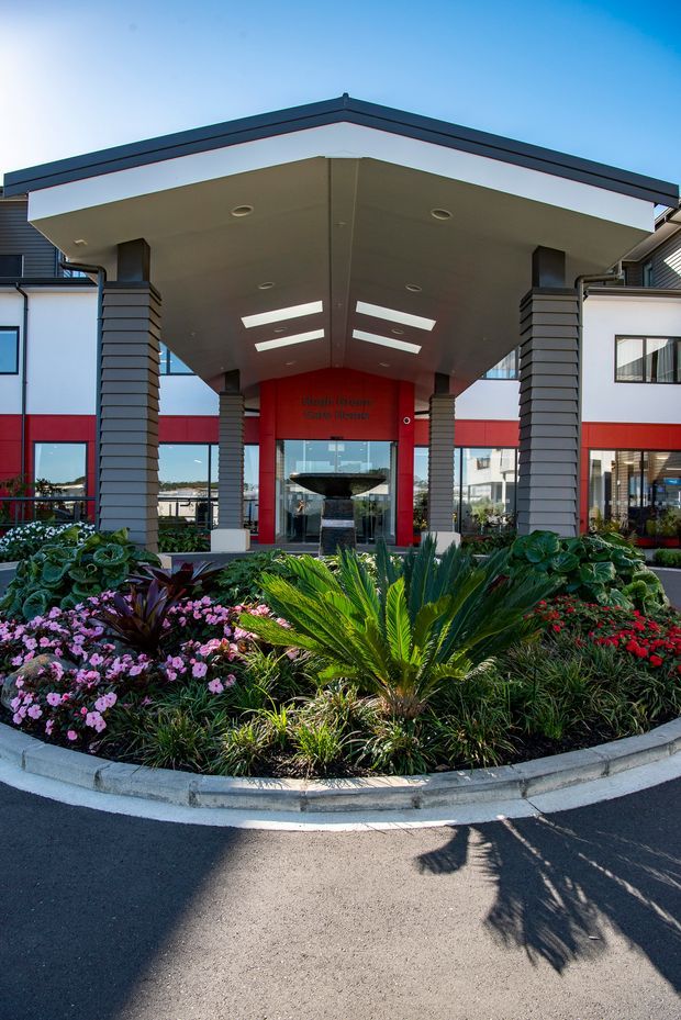 Front exterior view of a contemporary commercial building entrance with a large projecting canopy supported by dark horizontal-clad columns. The facade combines white walls, red accent panels, extensive glazing, and a landscaped circular driveway island with tropical and flowering planting.