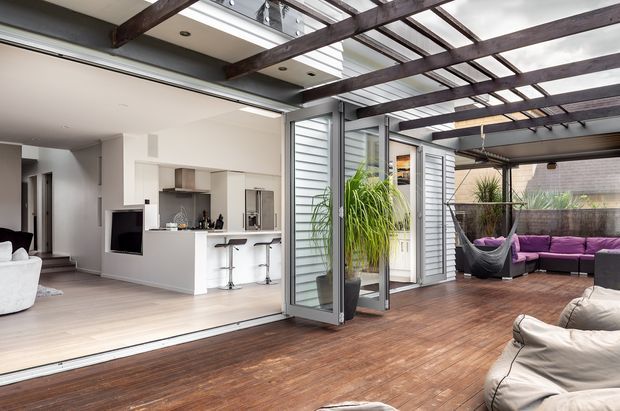 A contemporary indoor-outdoor living space with a large timber deck and covered pergola framed by dark exposed beams and translucent roof panels. Wide sliding glass doors open into a bright open-plan kitchen and living area with white cabinetry, a peninsula with bar stools, light timber flooring, and a wall-mounted television. The covered outdoor zone includes lounge seating, a hanging chair, and planters, creating a relaxed entertaining area.