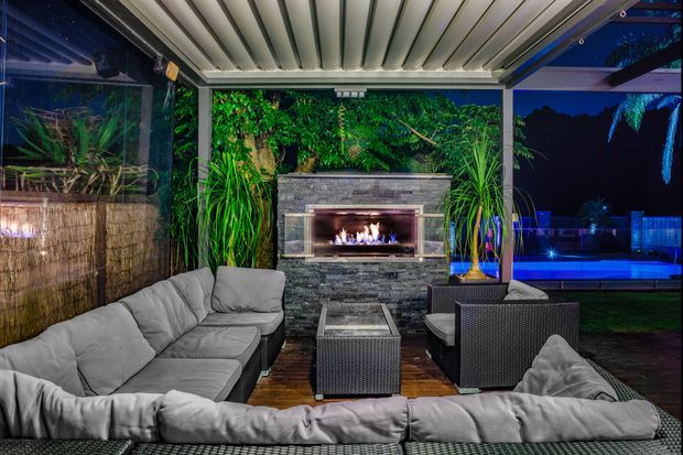Covered outdoor lounge at night with a large grey sectional sofa, woven armchairs, coffee table, and a stone-clad fireplace with an active fire. The space is surrounded by lush planting and overlooks a brightly lit swimming pool, with a slatted roof and screened side enclosure creating a sheltered entertaining area.