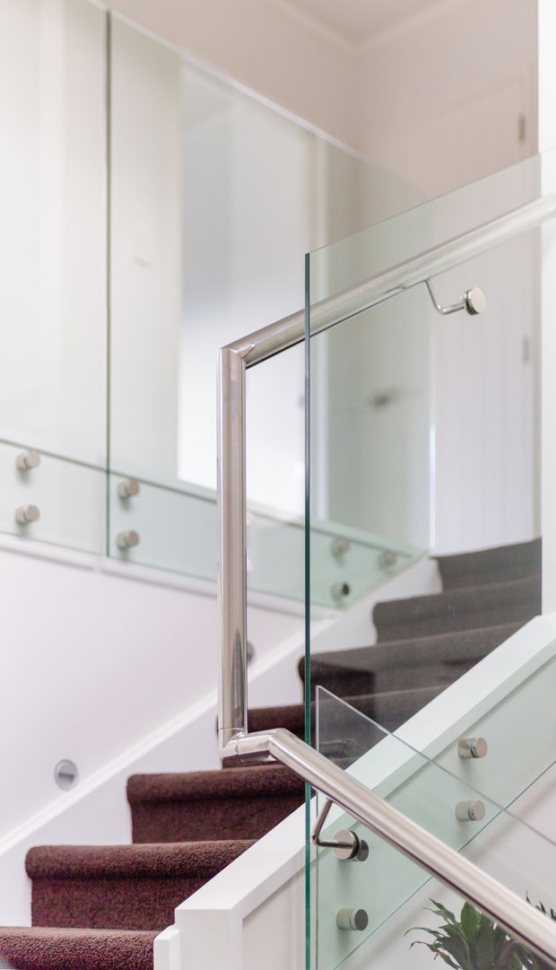A bright modern stairway with carpeted steps, white walls and balustrades, extensive clear glass panels, and polished metal handrails. Mirrored or reflective surfaces visually enlarge the space and reflect built-in cabinetry with small round handles. The composition focuses on the staircase and its contemporary glass-and-metal detailing.