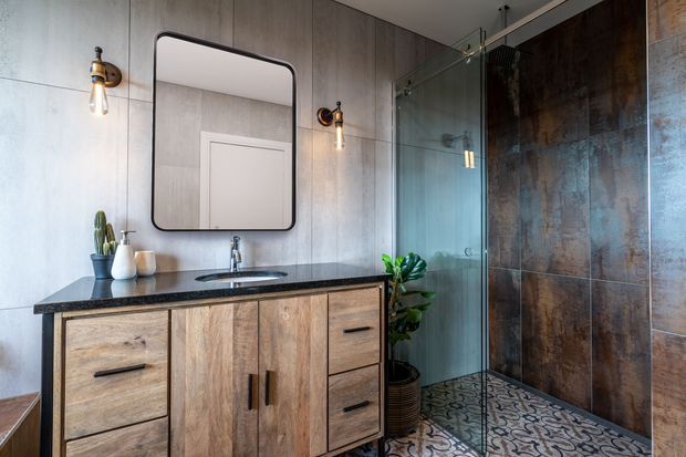 A contemporary bathroom vanity with a black stone benchtop, timber-look cabinetry, wall-mounted basin, rounded-corner mirror and exposed wall sconces. The bathroom includes a frameless glass shower enclosure with large weathered metal-look wall tiles and patterned encaustic-style floor tiles.