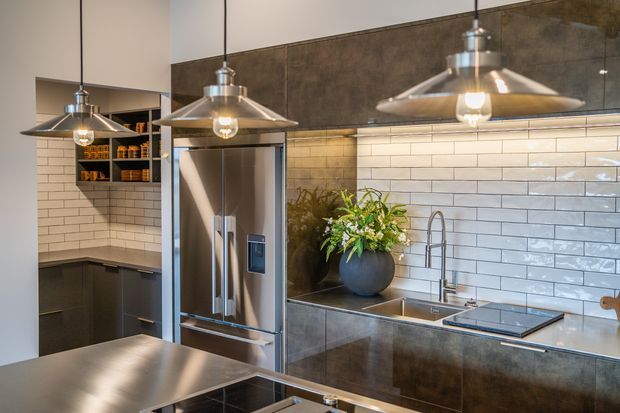 Contemporary kitchen featuring dark cabinetry, stainless-steel appliances and benchtops, white horizontal subway-tile splashbacks, and three industrial-style pendant lights. A concealed pantry or storage nook is visible at left, while a sink, cooktop, refrigerator, and decorative plant define the main kitchen workspace.