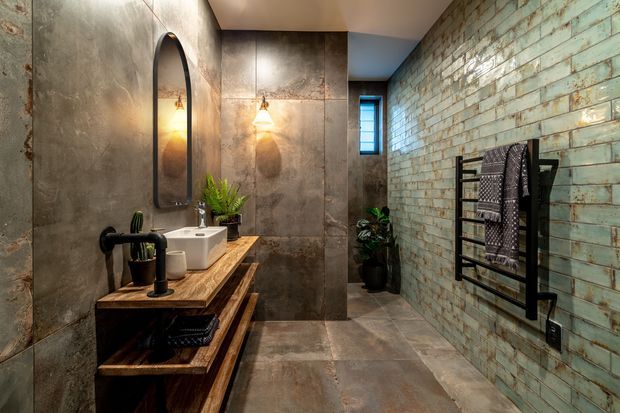 A moody contemporary bathroom with a timber vanity, white vessel basin, black tapware and fixtures, a tall arched mirror, and warm wall sconces. Large weathered metal-look wall panels contrast with a turquoise glazed brick feature wall, while a black heated towel rail and indoor plants add functional and natural accents.