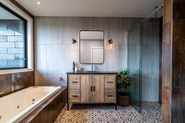 A stylish contemporary bathroom featuring a freestanding spa bath beside a large window, a timber-look double vanity with black benchtop, a frameless glass shower, patterned encaustic-style floor tiles, and warm timber wall and column finishes. Brass wall sconces and greenery add warmth to the refined space.