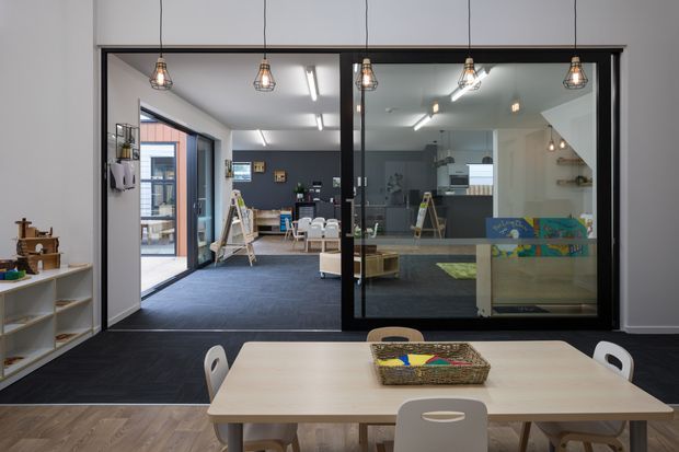 A contemporary multipurpose interior viewed from a light timber-floored dining or meeting area through black-framed glazed partitions. The foreground contains a large pale timber table with white chairs, while the adjoining carpeted room includes low shelving, display boards, artwork, pendant lights and fluorescent ceiling lighting. A glazed entry or window wall provides additional daylight, and a staircase with shelving is visible at the right.