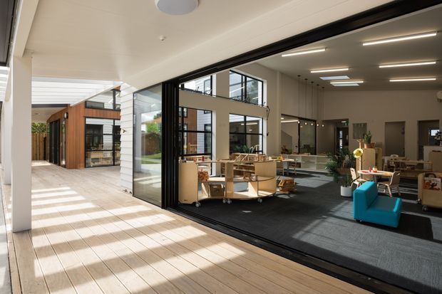 A bright contemporary commercial interior opening onto a covered timber-decked courtyard or walkway. Large black-framed glazed sliding doors reveal an open-plan office or communal workspace with desks, shelving, lounge seating and plants. The space features a high ceiling with linear lighting and skylights, while an adjacent timber-clad pavilion and landscaped courtyard are visible outside.