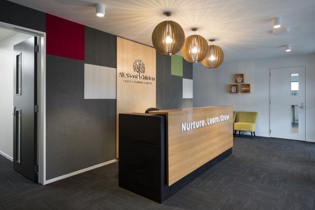 A contemporary reception area for a childcare centre, featuring a timber reception desk with black side panels, a colourful geometric feature wall with signage, dark patterned carpet, a white textured ceiling, and three oversized timber pendant lights. A yellow lounge chair and small wall-mounted display shelves add warmth to the otherwise professional entry space.