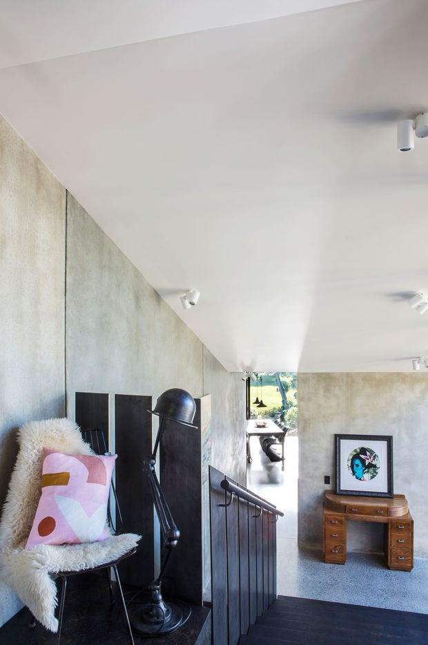 Interior view from a stairway or landing, looking down toward a bright open living area. The space has a dark timber staircase and balustrade, pale textured concrete-like walls, a white ceiling with track spotlights, and dark flooring. A sculptural black floor lamp, fur-covered chair with a colourful geometric cushion, and mid-century-style wooden drawer console with framed artwork add character.