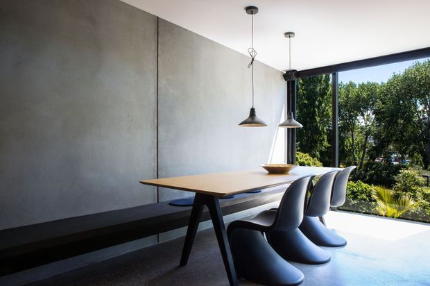 Contemporary dining area with a long timber dining table, sculptural dark chairs, and a built-in dark bench against a smooth grey concrete-look wall. Two suspended pendant lights hang above the table, while full-height corner glazing opens views to lush greenery outside. The room has a clean, minimalist composition with strong natural light and a refined material palette.