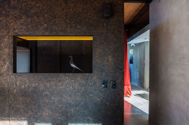A dark, dramatic interior view dominated by a textured charcoal-brown feature wall with a recessed horizontal opening or niche. The niche contains dark built-in panels, a bright yellow linear light, and a sculptural chair visible within it. A doorway at right leads to a brighter adjoining space with a vivid red curtain, concrete-toned walls, and timber ceiling detailing. The composition emphasises strong contrast between the moody feature wall and sunlit corridor.