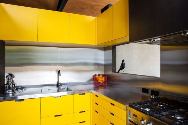 A compact, highly saturated yellow kitchen with L-shaped cabinetry, stainless-steel benchtops and splashback, integrated rangehood, sink, and freestanding gas cooker. The cabinetry reaches toward the ceiling, while a dark timber ceiling and black accents provide contrast. The space has a bold, utilitarian contemporary appearance.
