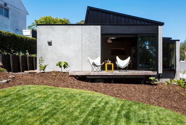 A compact contemporary backyard pavilion or studio with a dark mono-pitched roof, raw grey concrete or cement-finished wall, black-framed glazing, and a timber deck. The open front reveals a small seating area with two white sling chairs and a yellow side table. It sits within a landscaped suburban garden with lawn, mulched planting beds, hedges, and raised planters.
