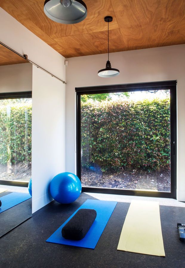 Bright, compact home gym or exercise studio with large black-framed windows overlooking a hedge. The room has dark flooring, blue and pale exercise mats, a blue stability ball, a black bolster, mirrored glazing on the left wall, and pendant and flush-mounted ceiling lights. The ceiling is clad in warm-toned timber with visible grain, while the walls are painted off-white.