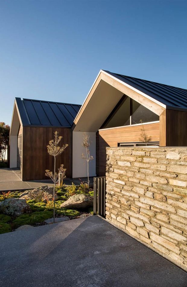 Exterior view of a contemporary residential property with two steeply pitched gabled forms, dark standing-seam metal roofs, warm timber cladding, large triangular glazing, and a textured natural-stone boundary wall. A landscaped entry garden and paved driveway lead toward the buildings under clear daylight.