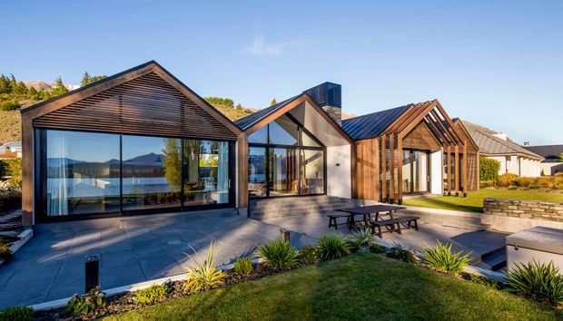 Exterior view of a contemporary multi-volume house with steep gabled roofs, extensive floor-to-ceiling glazing, dark metal roofing, and warm timber-lined gable façades. A paved terrace with outdoor dining furniture connects the building to a landscaped garden, with hills and water visible in the background.