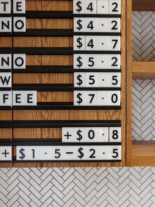 Close-up of a restaurant-style letterboard menu mounted against warm wood panelling, with black rails and white interchangeable letter and number tiles displaying prices. A white and light-grey herringbone tile wall and timber-framed shelving are visible beside and below the board.