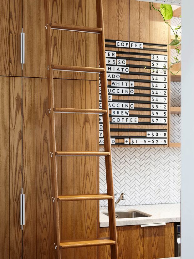 A warm, wood-clad café kitchen or service area featuring tall timber cabinetry, an integrated ladder, a letter-board coffee menu, white herringbone tile backsplash, and a stone countertop with sink. Greenery is visible near the upper right.