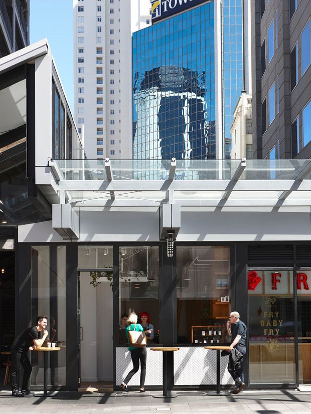 Street-level view of a contemporary urban commercial frontage with a café or retail tenancy beneath a glass-and-metal canopy. The facade uses extensive blue reflective glazing, dark framed windows, white structural elements, and a transparent glass balustrade, with surrounding high-rise buildings visible in the background.
