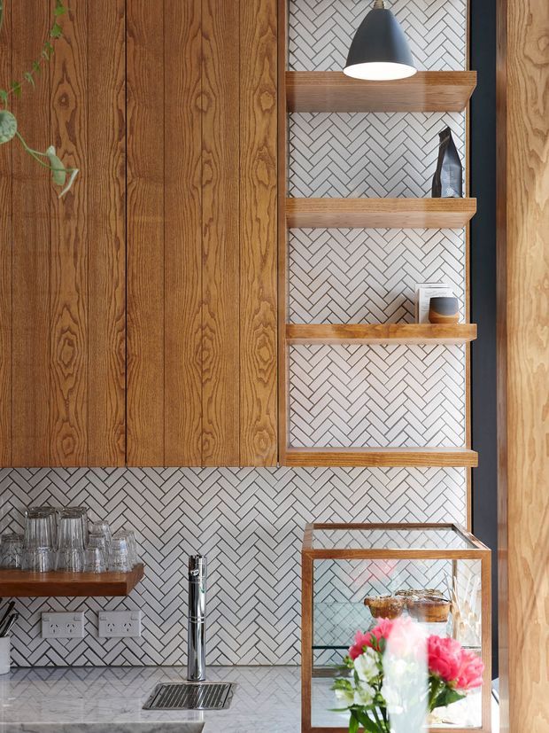 Close-up view of a refined kitchen or beverage station featuring warm vertical timber cabinetry, open timber display shelves, a white and grey herringbone tile backsplash, a marble-look benchtop, chrome tapware, and a glass-front display cabinet. A black pendant light and small decorative objects complete the composition.