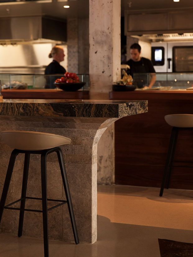 A warmly lit commercial kitchen or hospitality bar interior, viewed across a stone-clad counter with high stools. The foreground counter has a sculptural, angled stone base and a dark patterned stone benchtop. Behind it are timber cabinetry, stainless-steel kitchen equipment, a glass display or partition, staff members, and a serving counter. The atmosphere is refined, earthy, and intimate, with concrete and timber architectural elements.