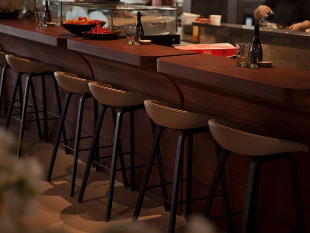 A dimly lit hospitality bar or counter with a long dark timber-clad benchtop, several tan upholstered bar stools with black metal legs, glassware, bottles, and food displays. The warm, intimate composition focuses on the bar seating and built-in counter, with a staff member partially visible behind the service area.